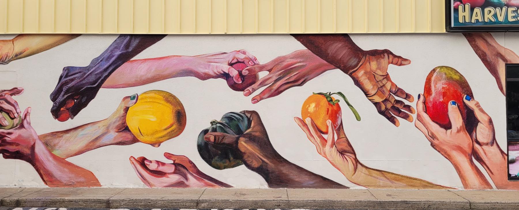 A mural on an external wall of a grocery store; on a white background,  the mural depicts hands of various skin tones handing pieces of fruit vegetables to others. "No Parking" is painted on the pavement in front of the mural.