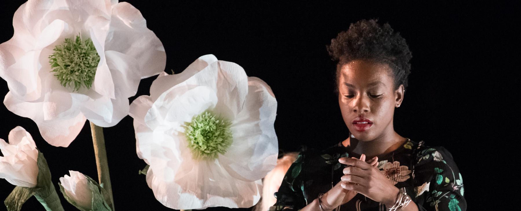 On a stage with spotlights, Deborah crosses her arms next to very large, white daisies. 