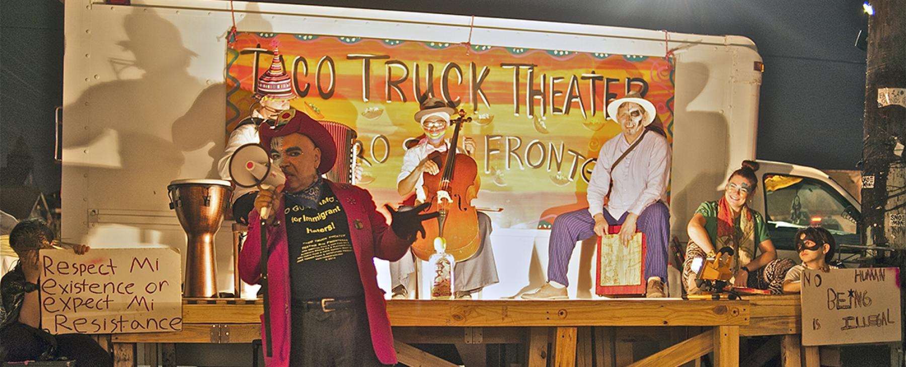 In front of a "Taco Truck Theater" sign, three musicians perform on a stage. In front of them, a man speaks into a mic, while wearing a red cowboy hat and jacket.