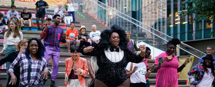 A Black drag queen, with a big afro and tuxedo body suit, leads a group in a dance.
