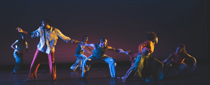 Indoors, on a stage, four performers hold hands forming a chain. There are two people behind them mid movement, and a woman in front.