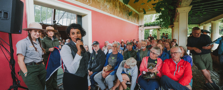 A woman dressed in a denim vest, a button-up shirt and dark pants speaks into a microphone while a seated audience watches.