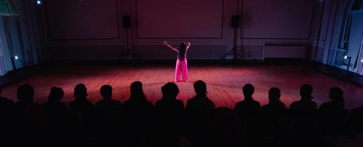 The dark silhouette of a row of seated audience members makes up the bottom half of the image. Further up, a woman in bright pink pants and a pink top  stretches out an arm, her back to the audience.
