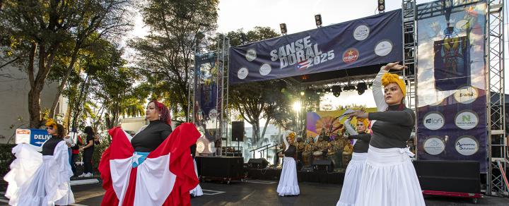 Outdoors and in daylight, four women in long gowns and colorful headwraps dance.
