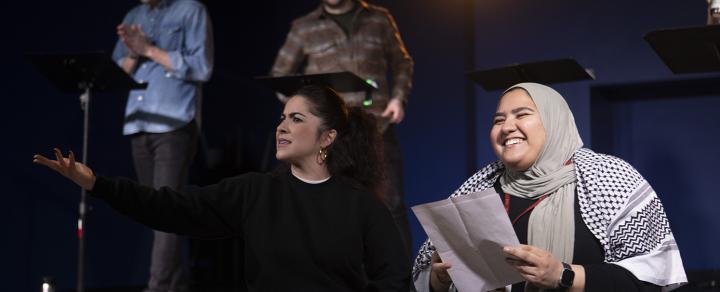 Two women of color, one in a hijab, sit on the edge of a stage. Both hold scripts. More folks are behind them in the shadows.