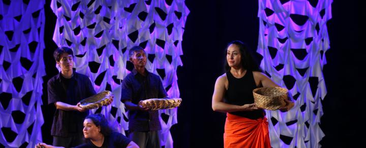 Three performers in all black, hold baskets and one kneels. They are behind a woman who wears a sarong and holds a basket.