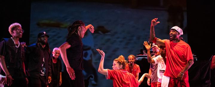 A group of predominantly Black folks form a dance circle. Three folks are in the middle bending their arms and dropping it low.
