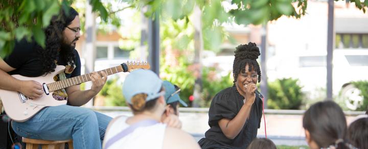 A Black woman sings into a microphone next to a guitarist. They're outside and in front of kids with chaperones.