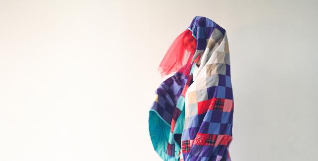 Under a multi-color quilt, a person peaks out. They pose in front of a white backdrop.