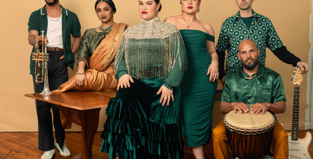 A group of six dressed in green pose in front of a mustard colored wall.