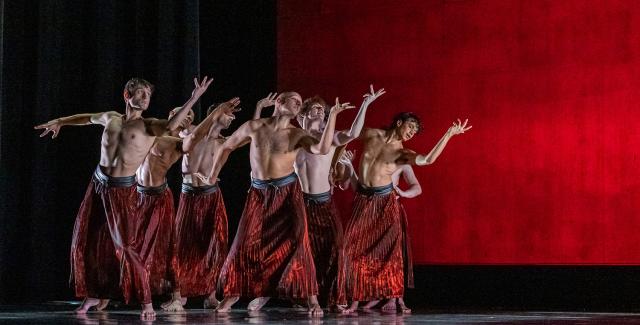 On a stage, in front of a red backdrop, seven dancers in only red pants hold their left hands up and their right arm to their sides.