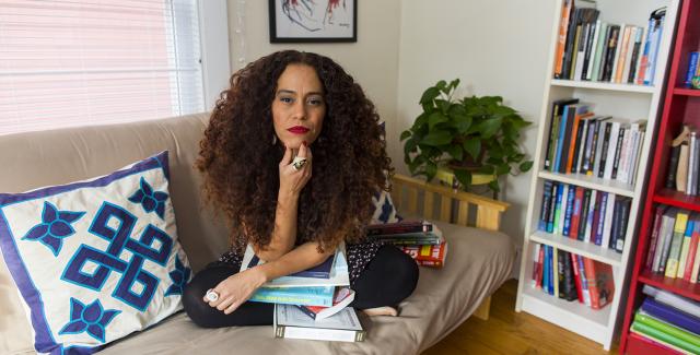 Yara makes eye contact with the camera. She leans over books on her lap in her living room.