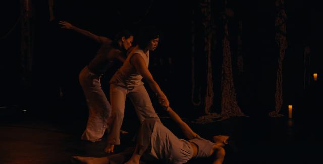 In the dark, two dancers help another get off the stage. They wear the same color tank tops and pants of different cuts.