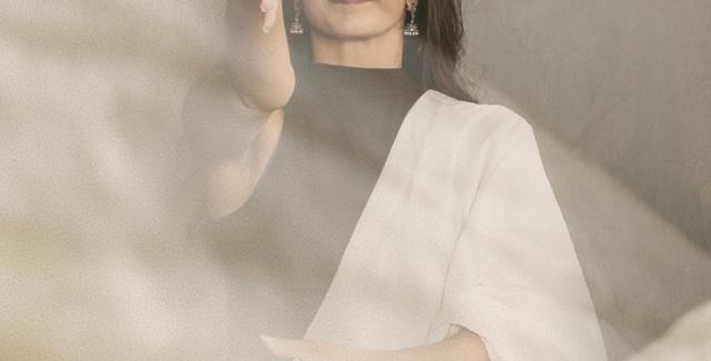 An Indian woman is behind a sheet fabric in the foreground. She holds her hand out and wears traditional garb.