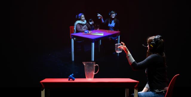 A person at a table holding a glass looking at a projection of  two people at another table holding glasses.