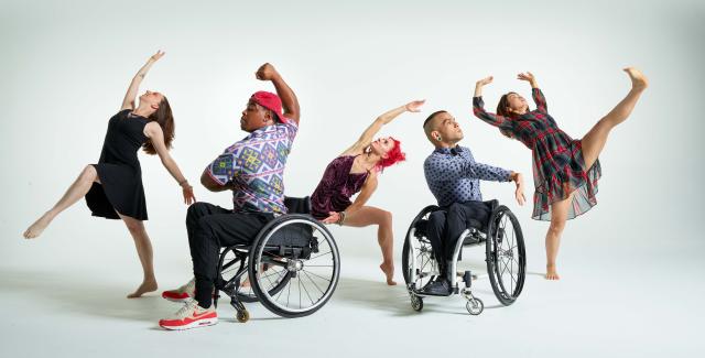 In front of a white backdrop, five dancers (two in wheelchairs) perform with their arms or legs out wide.