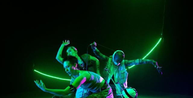 Four dangers, with their arms out and a couple of them in headpieces like helmets, perform with their arms wide and in front of a curved neon green light.