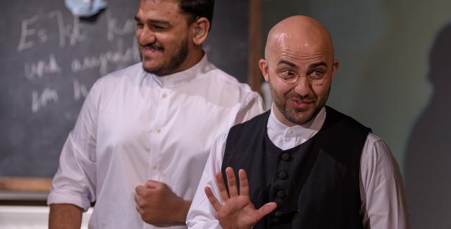 Brown men in white shirts stand in front of a chalk board in stage lighting.