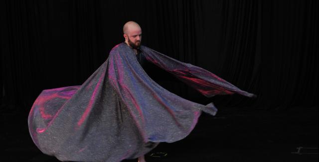 A dancer on a dark stage wearing a large cape which is swirling around. Only the dancer's head and feet are visible.