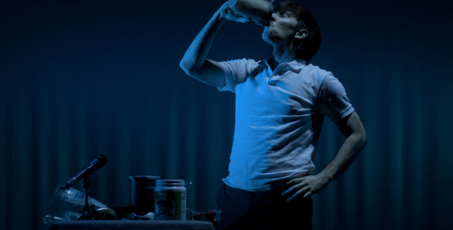 A person in a white polo shirt drinks from a mug in front of a dimly lit curtain.