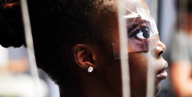 A young black girl wears a mask taped over her eyes of bigger eyes.