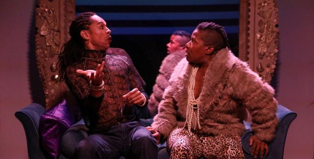 Two Black folk sit on a couch with stage lighting. They gesture to each other.