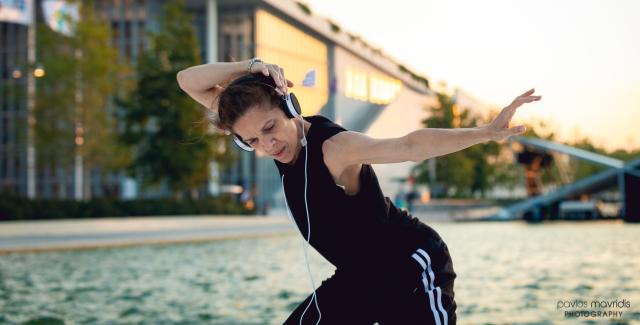 Outside, on the beach, Ana dances with a hand holding a headphone to her ear. She is in all black, including a sleeveless tee.