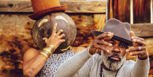 A Black woman holds a mask over her face, while wearing a top hat, and, in front of her, a Black man, in a fedora, holds his hands up.