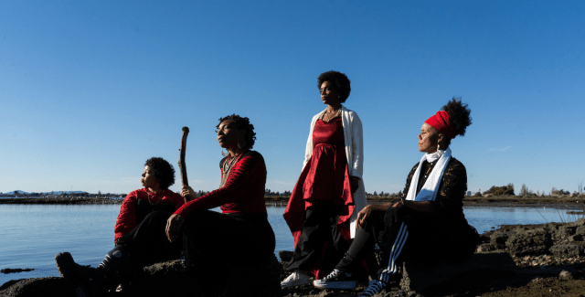 Four people are outside by a waterfront, three of them sit and one of them stands.