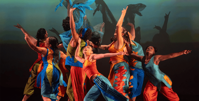 On a stage, folks in blues and oranges spread their arms and some hold blue wings above their heads.