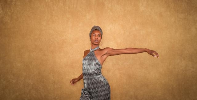 In front of a tan backdrop, a Black woman, in a grey headwrap and matching satin jumpsuit, holds her arm out to her left.