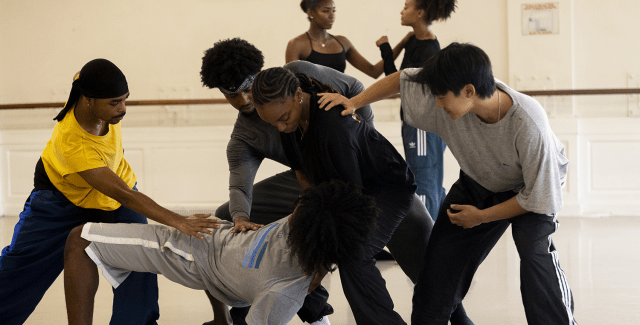 In athleisure and in a studio space, five performers hold another who leans almost all the way down to the ground. 