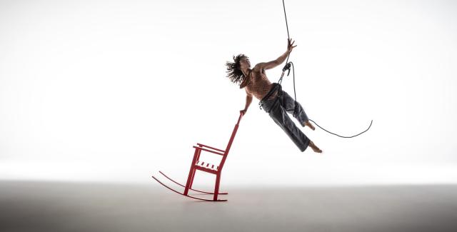 A person suspended in midair by a harness reaches upward with their left hand while touching a red rocking chair with their right.