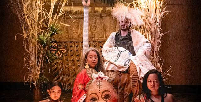 Four performers sit together under and behind neon lights. Three women hold large masks and a man sits behind them in a garment with wide and long sleeves.