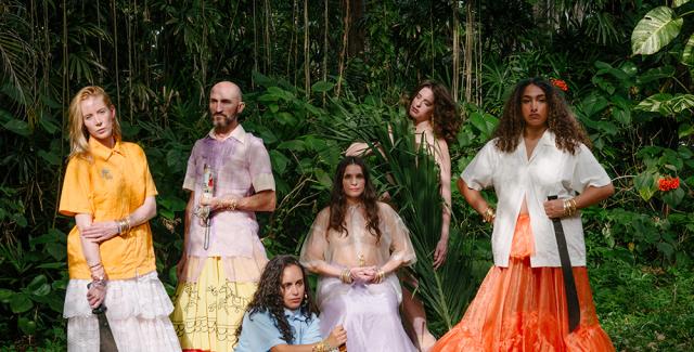 7 dancers wearing guayaberas and long skirts are arranged kneeling, laying, crouching, and sitting in a tropical outdoor setting. They look into the camera with varying expressions of intensity and devotion. Some carry machetes or other symbols of cuban American iconography.