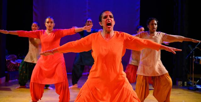 A brown woman in orange dress holds her arms out and screams. Four dancers are behind her.