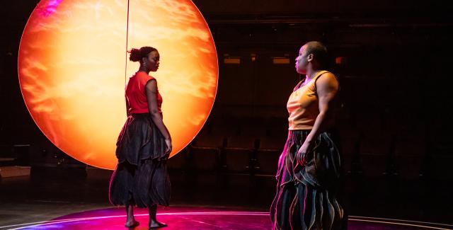 In front of a sun-like orb on a stage, two Black women dance in warm tank tops and billowing grey skirts.