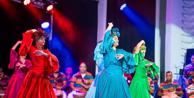 On a stage, four women in monochromatic but bright colored costumes perform. They wear headwraps that match their dresses.