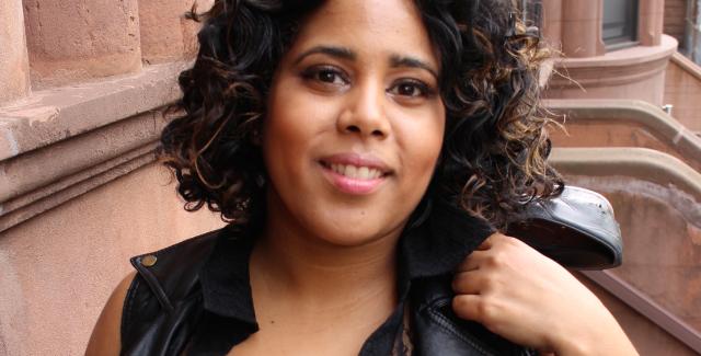 A brown woman holds shoes over her shoulder and has curly dark hair.