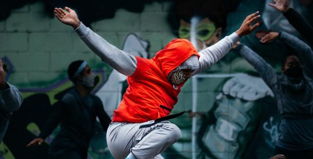 On a street and in front of a mural, a dancer throws their arms and leg up while wearing an orange hoodie.