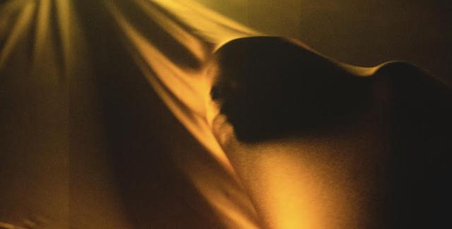 Under a spotlight, a person spreads golden satin over their face and stretches it with their hands.