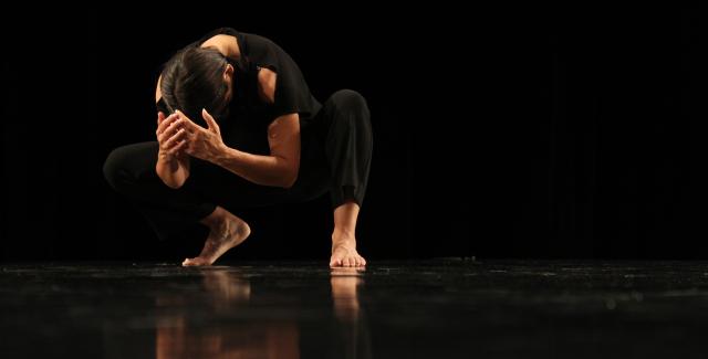 In a black background with body angled frontward and slightly towards the left, a Native woman dancer with medium brown skin, dark brown straight hair crouches in a semi-squat position with a curved back, elbows bent, and criss-crossed hands slightly covering the top of her head facing downward. One foot is flat on the ground while the other’s heel is raised. She wears black pants and a sleeveless top exposing both arms and shoulders. The black shiny flooring reveals a blurred reflection of her body.
