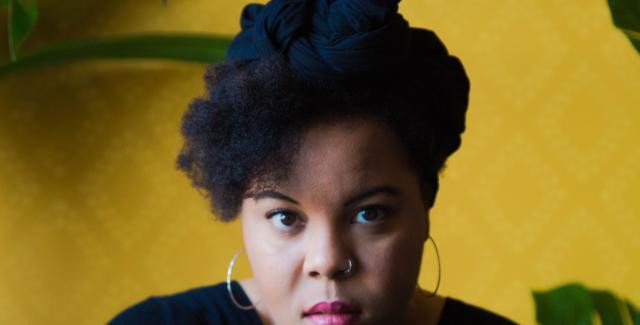 A Black woman poses in front of a yellow wall and bright green plants. She leans on her hand and wears gold hoops and all black.