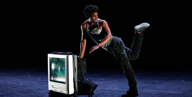 A performer kicks up a foot while leaning against a television box on a dark stage.
