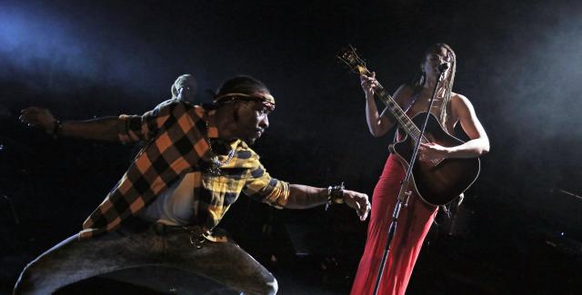 A Black woman in a red dress playing guitar and sings into a mic while a Black man dressed in jeans and yellow flannel appears to dance with arms outstretched and in a deep plie.