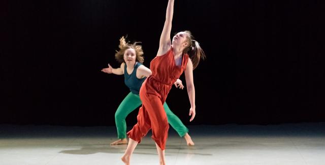 Two dancers, one in a rust jumpsuit with right arm extended straight up and another behind wearing shades of green with both arms outstretched