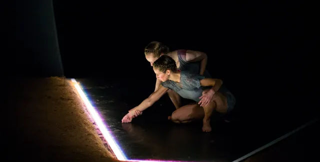Two performers crouch by a bright tube of light and inspect it.