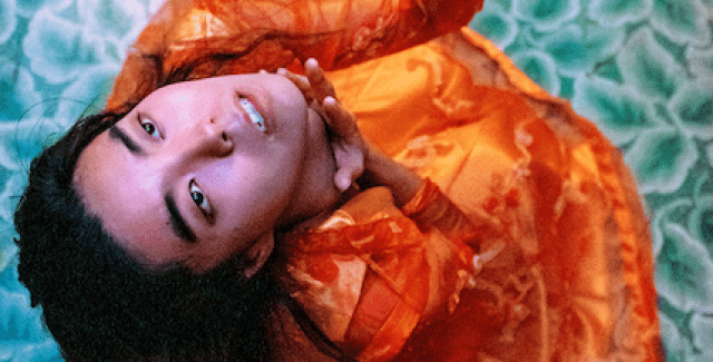 From above, a person dressed in orange is shown looking upwards while standing on a floral patterned blue floor.