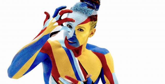 In a white space and covered in body paint, a person with a top bun touches their forehead.