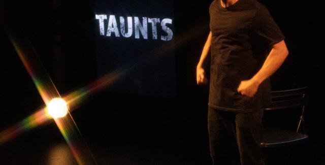 On a stage, a man of color stands before the word "Taunts." He is in all black and the stage is dimly lit.